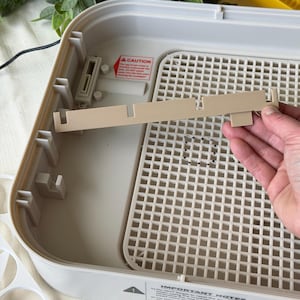 May include: A beige plastic incubator with a white interior and a removable grid. A beige plastic piece is being held above the incubator. The image also shows a warning label and a set of egg holders.