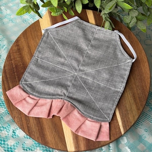 Puede incluir: Mascarilla gris con costuras blancas y un bajo rosa con volantes. La mascarilla tiene presillas elásticas para las orejas y se muestra sobre una tabla de cortar de madera.