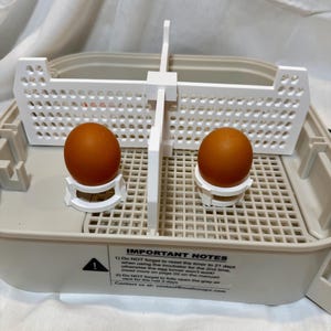 May include: An incubator with two brown eggs in white holders. The incubator has a white grid and a white divider. The text "IMPORTANT NOTES" is visible, with instructions about resetting the timer and opening the air vent.