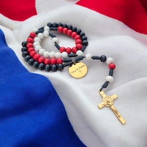 May include: A rosary featuring dark blue, white, and red beads strung on dark cord. A gold-toned metal crucifix hangs from the end, and a circular gold-toned metal charm is attached, engraved with the text Vive la France and a small heart symbol.