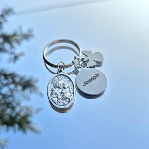 May include: Silver keyring with three charms: an oval pendant depicting St. Joseph and the child Jesus, a round charm with the name "Joseph", and a clover-shaped charm. The keyring is on a reflective surface.