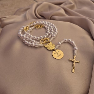 Può includere: Un rosario di perle con dettagli dorati. Il rosario presenta una croce dorata, un ciondolo rotondo con incisa la scritta "Nicole & Kevin" e un elemento decorativo con il testo "Two Souls One Heart".
