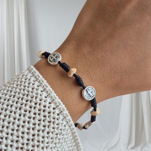May include: Black cord bracelet featuring silver-toned circular charms and light-colored beads. The charms have religious symbols. The bracelet is worn on a wrist, with a cream-colored sweater in the background.