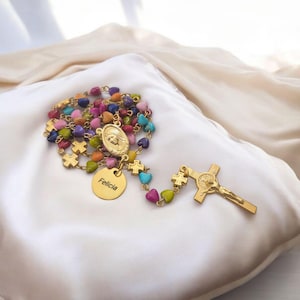May include: A gold-colored rosary with colorful heart-shaped beads. The rosary features a crucifix, a medallion with a religious image, and a round tag with the name "Felicia". The beads are in various colors, including pink, blue, and yellow.