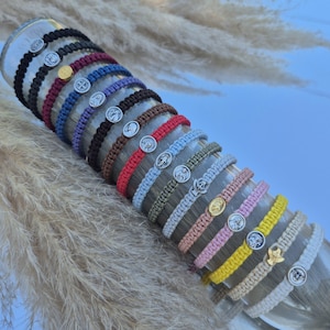 Puede incluir: Una colección de pulseras de macramé en varios colores, cada una con un medallón religioso. Las pulseras se exhiben en un cilindro de vidrio transparente, mostrando los diferentes colores de los cordones y los dijes de metal. Las pulseras son de color negro, rojo, azul y otros.