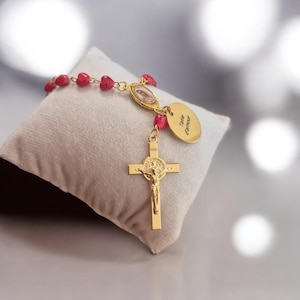 Può includere: Rosario dorato con perline rosse a forma di cuore. Il rosario presenta un crocifisso con una dettagliata raffigurazione di Gesù e un ciondolo ovale con la scritta "Tatie d'amour". Il rosario è esposto su un cuscino bianco.