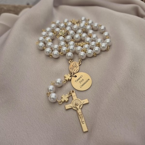 May include: A gold rosary with white pearl beads. The rosary features a cross pendant and a round charm engraved with "Tamara's First Communion." The rosary is displayed on a neutral fabric.