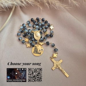 May include: A rosary featuring grey and black speckled beads and gold-coloured details. It includes a crucifix, cross-shaped connectors, and a medallion with a religious figure. The text "Choose your song" is displayed with a QR code.