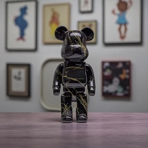 Puede incluir: Una figura negra con forma de oso con vetas doradas. El juguete coleccionable es brillante y se mantiene erguido sobre una superficie de madera. El fondo presenta obras de arte enmarcadas.