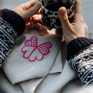 May include: A black and white speckled ceramic mug filled with a dark liquid, held by hands wearing a patterned sweater. A folded linen napkin with a pink embroidered floral design rests below the mug.