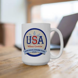 May include: White ceramic mug with a large handle. The mug features a circular design with the word "USA" in red and blue, along with "Milano Cortina 2026" and "Living Room Judging Panel" in blue text. The mug is on a wooden surface.