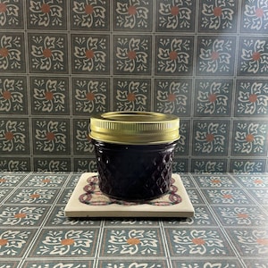 Puede incluir: Un pequeño tarro de mermelada morada oscura con tapa dorada se encuentra sobre un posavasos cuadrado decorativo. El tarro tiene un patrón texturizado. El fondo presenta un diseño de azulejos florales repetidos en tonos de verde azulado, naranja y blanco.