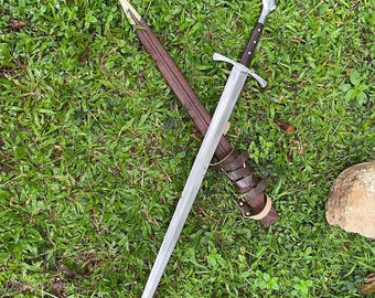 Puede incluir: Una espada plateada con una empuñadura en forma de cruz descansa sobre hierba verde junto a una vaina de cuero marrón. La espada tiene una hoja larga y afilada y un mango de madera oscura. La vaina está asegurada con correas de cuero.