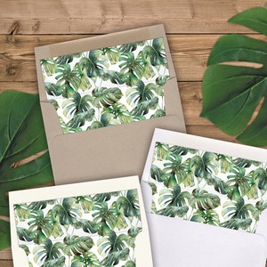 May include: Two white envelopes with a green and white tropical leaf pattern on the flap. The envelopes are on a wood background.