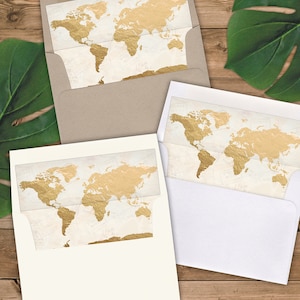 May include: Two white envelopes with gold foil world map designs. The envelopes are on a woodgrain surface with a green leaf in the background. The text "Click all photos for details!" is on a gold foil sticker.