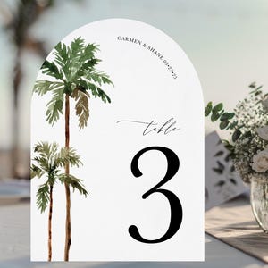 May include: White arch-shaped table number card with a tropical theme. The card features two palm trees with green leaves and brown trunks. The text "Carmen & Shane 03.23.23" is printed in the top right corner. The text "table 3" is printed in the center of the card.