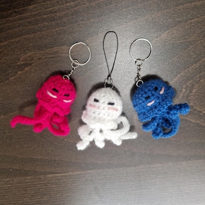 Peut inclure: Trois porte-clés poulpes crochetés à la main, aux couleurs vives : rose vif, blanc et bleu. Chaque poulpe a un visage souriant et est attaché à un porte-clés en métal ou à un cordon noir. Les porte-clés sont présentés sur une surface en bois.