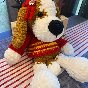 May include: A handmade, plush dog toy with a red and gold sweater. The dog has brown and white fur, a black nose, and a red bow with a logo. The toy is sitting on a striped tablecloth.
