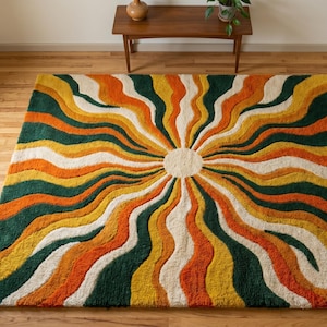 May include: A square area rug featuring a central cream-coloured circle with radiating wavy stripes in alternating shades of dark green, bright orange, and golden yellow, creating a sunburst pattern across the entire surface of the textile.