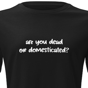 Puede incluir: Camiseta negra con el texto blanco "are you dead or domesticated?" La camiseta tiene mangas cortas y cuello redondo. El texto está en una fuente manuscrita.