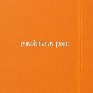 Op de afbeelding: Een fel oranje notitieboek met de tekst "mischievous pixie" in witte letters. Het heeft een bijpassende oranje elastische bandsluiting en een lint bladwijzer. De omslag is effen en helder oranje.