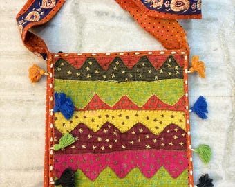Pode incluir: Bolsa de ombro artesanal com um design de patchwork colorido. A bolsa apresenta uma mistura vibrante de cores, incluindo laranja, verde, amarelo e rosa, com borlas decorativas e uma alça estampada. A bolsa tem aproximadamente 30 cm de largura e 36 cm de altura.