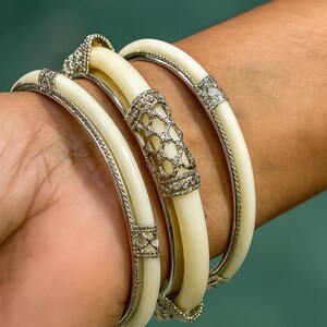 May include: Ivory-colored bangles with silver-toned accents and sparkling embellishments. The bracelets feature a central decorative element with a lattice design and are set with small, clear gemstones. The jewelry is displayed on a person's wrist.