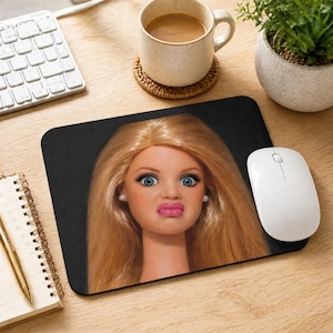 Può includere: Un tappetino per mouse rettangolare nero con un primo piano del viso di una bambola con capelli biondi, occhi blu e un'espressione sorpresa. Un mouse per computer bianco è a destra del tappetino. Sono visibili anche una tazza di caffè, una tastiera e un blocco note.