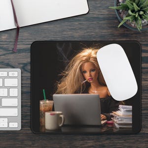 Op de afbeelding: Zwarte muismat met een pop met lang blond haar, die een sigaret rookt terwijl ze op een laptop werkt. Een witte muis, een koffiekopje en een ijsdrankje staan ook op de mat.