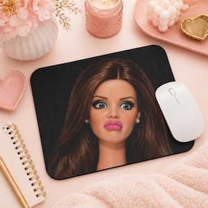 May include: Black rectangular mousepad featuring a doll's face with a surprised expression, large eyes, and pink lipstick. A white wireless mouse is on the right. The mousepad is on a pink surface with other decorative items.
