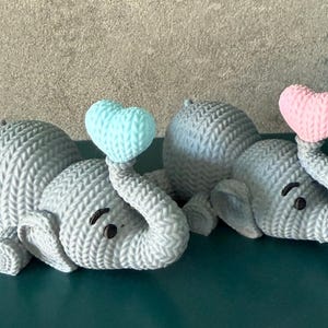 Può includere: Due statuette di elefanti grigi con corpi strutturati e proboscide sollevata che tengono palloncini a forma di cuore. Un cuore è azzurro, l'altro è rosa. Gli elefanti sono sdraiati su una superficie turchese.