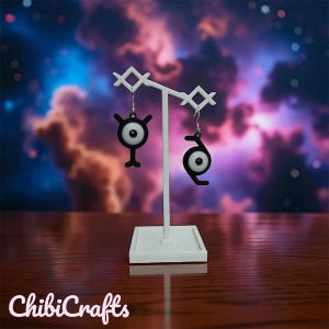 Könnte beinhalten: Ein Paar schwarz-weiße Ohrringe im Unown-Design, präsentiert auf einem weißen Ständer. Die Ohrringe haben ein stilisiertes Design mit schwarzer Umrandung und einem weißen, augenartigen Zentrum. Der Hintergrund ist eine farbenfrohe Mischung aus Blau, Rosa und Lila.
