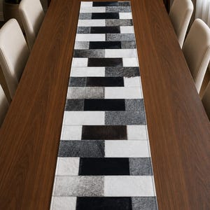 May include: A rectangular table runner featuring a patchwork design of rectangular panels in shades of black, white, and gray. The runner is constructed from individual pieces of textured material arranged in a repeating geometric pattern.