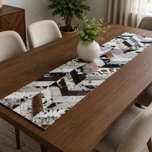 May include: A rectangular table runner featuring a chevron pattern made of cowhide patches in shades of brown, black, white, and grey. The runner is laid flat on a dark brown wooden dining table, centered beneath a white ceramic vase with greenery.