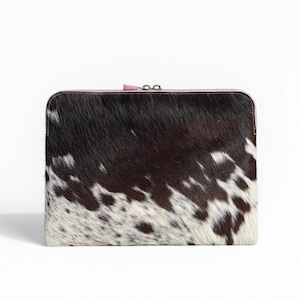 May include: A rectangular laptop sleeve with a cowhide pattern. The sleeve features a mix of black, brown, and white patches. It has a pink zipper and trim. The overall design is a blend of rustic and modern aesthetics.
