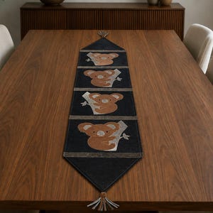 May include: A long, rectangular dark navy blue table runner featuring four repeating embroidered patches of brown koalas clinging to light gray tree branches. The runner has pointed ends finished with small, decorative gray fabric tassels.
