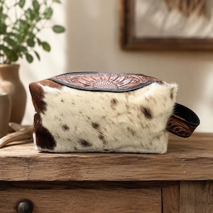 Op de afbeelding: Een rechthoekige toilettas met een lichaam van wit en bruin koeienhuid, voorzien van een donkerbruin lederen bovenpaneel en een zijhandvat. De lederen delen zijn versierd met een in reliëf aangebracht bloemmotief en de tas heeft een ritssluiting.