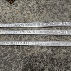 May include: Three silver metal bars with rounded ends, each stamped with the text "NATIONAL LEAD CO." and other markings, are arranged horizontally. The bars are lying on a mottled grey surface. The bars appear to be made of lead.