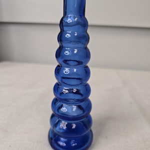 May include: A cobalt blue glass bud vase with a unique, stacked bubble design. The vase has a narrow neck and a rounded base. The glass is transparent, allowing light to pass through. This decorative vase is perfect for displaying a single flower or small floral arrangement.