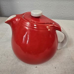 Puede incluir: Una tetera de cerámica roja con asa, boquilla y tapa blancas. La tetera tiene un cuerpo redondeado y un acabado brillante. La tapa tiene un pequeño pomo blanco redondo. La tetera se utiliza probablemente para preparar té.