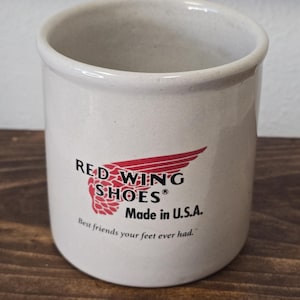 Könnte beinhalten: Beige Keramikbecher mit dem Red Wing Shoes Logo in Rot und Schwarz. Der Text "Made in U.S.A." und der Slogan "Best friends your feet ever had." sind ebenfalls auf dem Becher aufgedruckt.