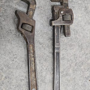 May include: Two vintage adjustable pipe wrenches. The wrenches are made of metal and show signs of wear. The jaws are open, ready for use. One wrench has a curved handle, while the other has a straight handle. The tools are on a gray surface.