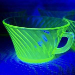 Op de afbeelding: Een set vintage groene glazen theekopjes met een spiraalvormig ontwerp. De kopjes hebben een oor en staan op een blauw oppervlak. Het glas gloeit met een neon groene tint, wat een opvallend visueel effect creëert. De kopjes zijn waarschijnlijk uit het midden van de eeuw.