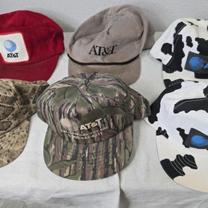 May include: A collection of six baseball caps with the AT&T logo. The caps are in various colors and patterns, including red, tan, camouflage, and a black and white cow print. Some caps have a patch with the AT&T logo.