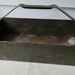 Puede incluir: Un portaherramientas de metal verde oliva con asa. El portaherramientas rectangular tiene un asa que se arquea hacia arriba. El metal muestra signos de desgaste y envejecimiento, con algo de óxido y decoloración.