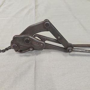 May include: A vintage, rusty metal cable grip tool with a chain attached. The tool has a hinged design with a curved handle and is likely used for pulling or securing cables. The tool is set against a white background.