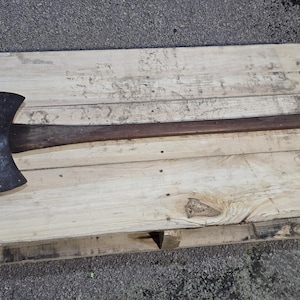 Puede incluir: Un hacha de doble filo con una cabeza de acero oscura y desgastada y un mango largo de madera. El hacha descansa sobre un palet de madera de color claro, mostrando su diseño rústico. Es una herramienta para cortar madera.