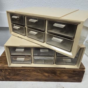 May include: Two beige plastic storage organizers with clear drawers. Each organizer has two rows of four drawers. The organizers are stacked on top of a dark brown wooden box.