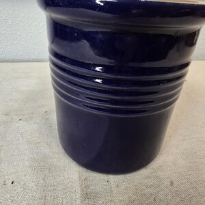 Może przedstawiać: Głęboki niebieski ceramiczny doniczka z błyszczącym wykończeniem. Doniczka ma cylindryczny kształt z poziomymi żebrowanymi detalami wokół korpusu. Obręcz jest lekko rozszerzona, a wnętrze ma jaśniejszy kolor. Ta dekoracyjna doniczka nadaje się do użytku wewnątrz i na zewnątrz.