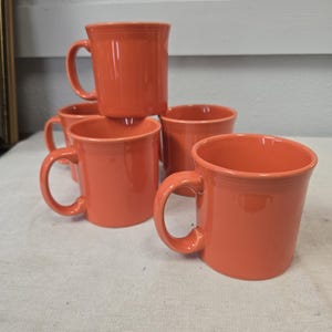 Puede incluir: Una pila de cinco tazas de cerámica naranja con asas redondeadas. Las tazas son de un color naranja vibrante y sólido y parecen brillantes. Las tazas están dispuestas sobre una superficie blanca. Cada taza tiene una capacidad de unos 350 ml.
